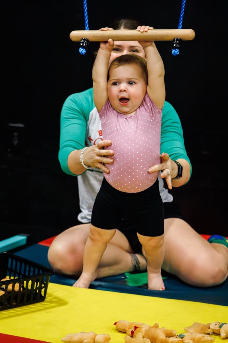 A coach holding a toddler up to a wooden bar in a Baby Sharks class