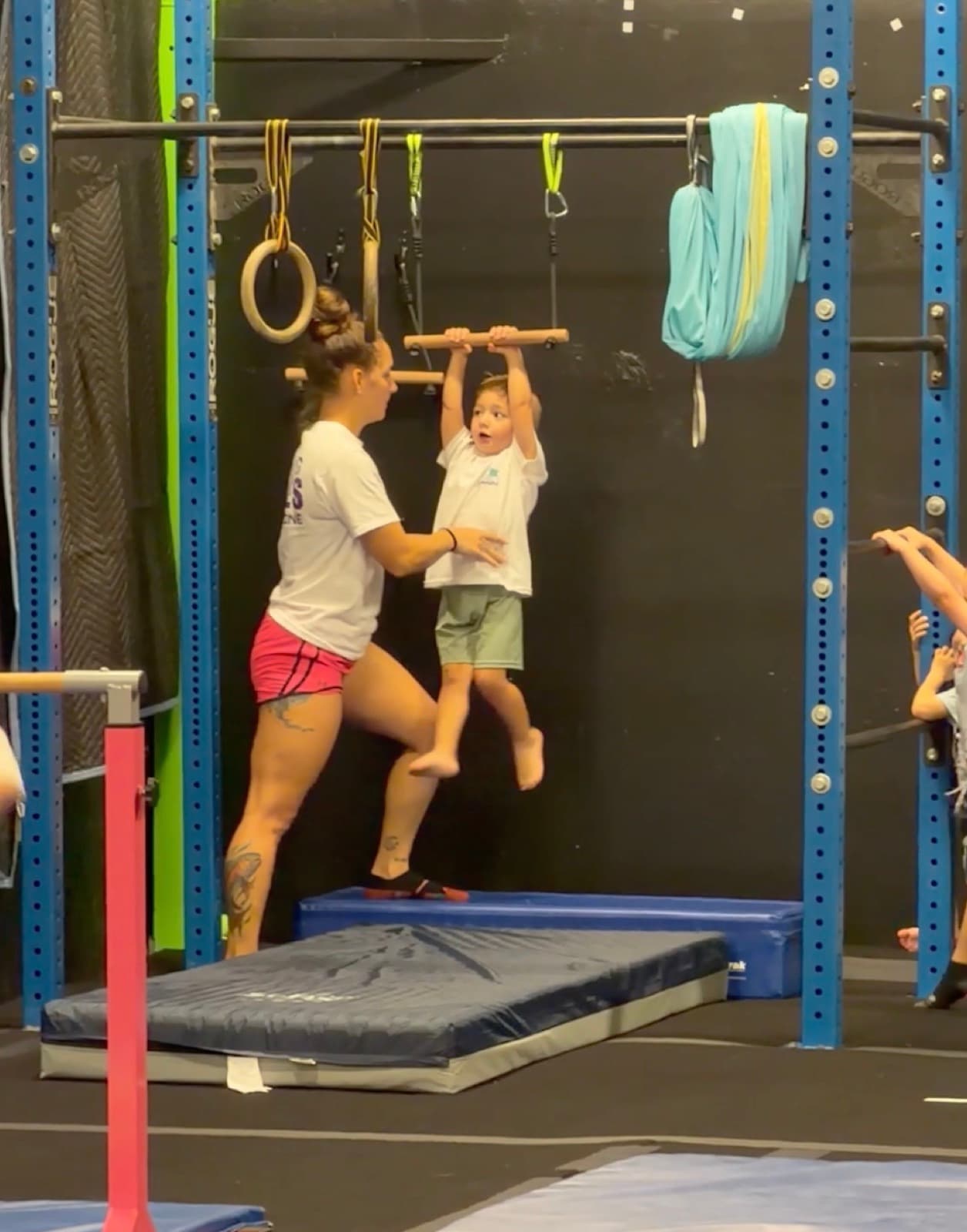 An older child hanging from the trapeze bar while a coach spots